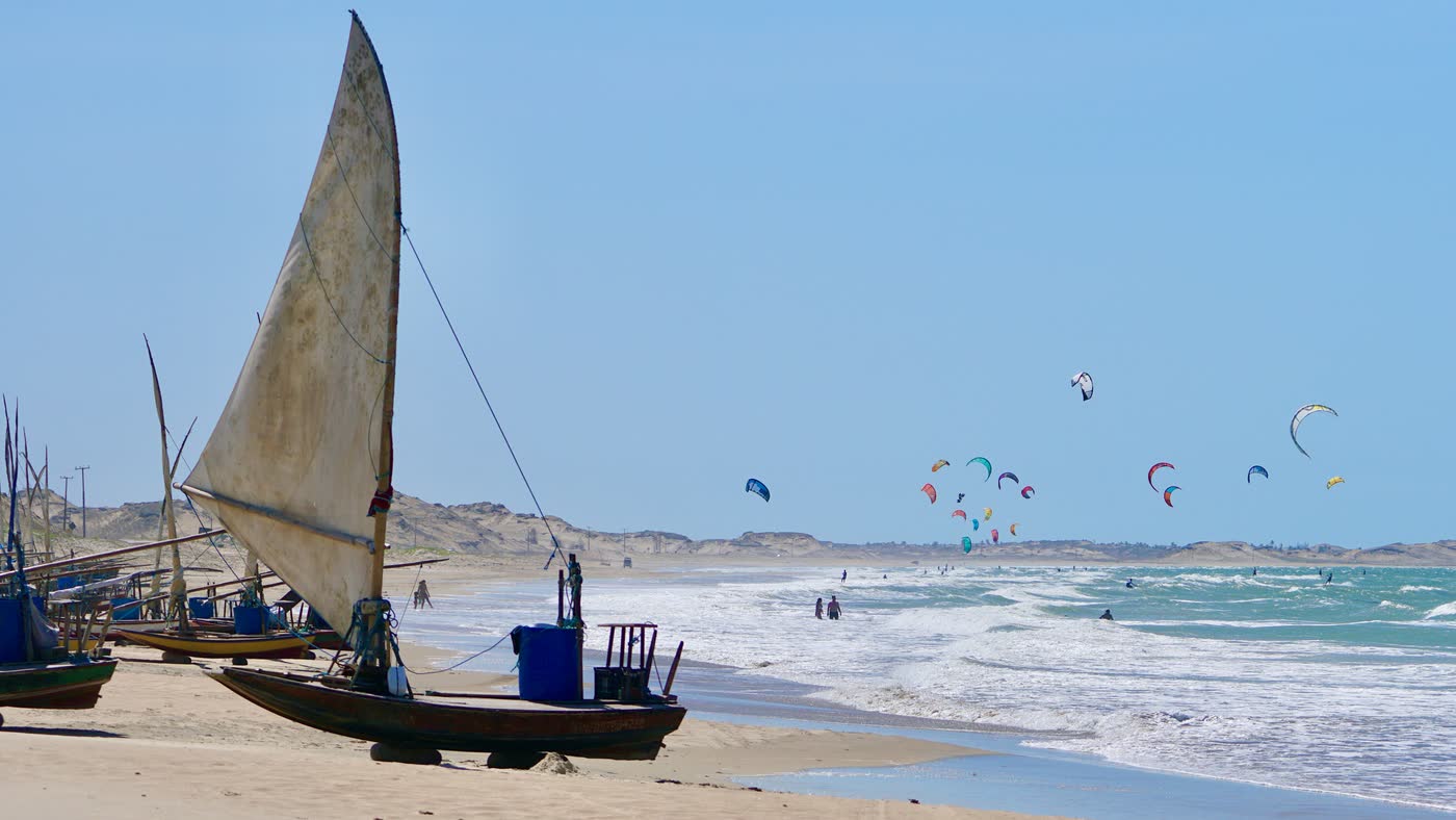 Jangada na areia com dezenas de kites no ar ao fundo, no litoral do Ceará